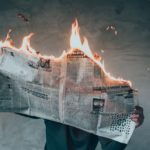 A man reads a newspaper that is on fire while standing against a wall