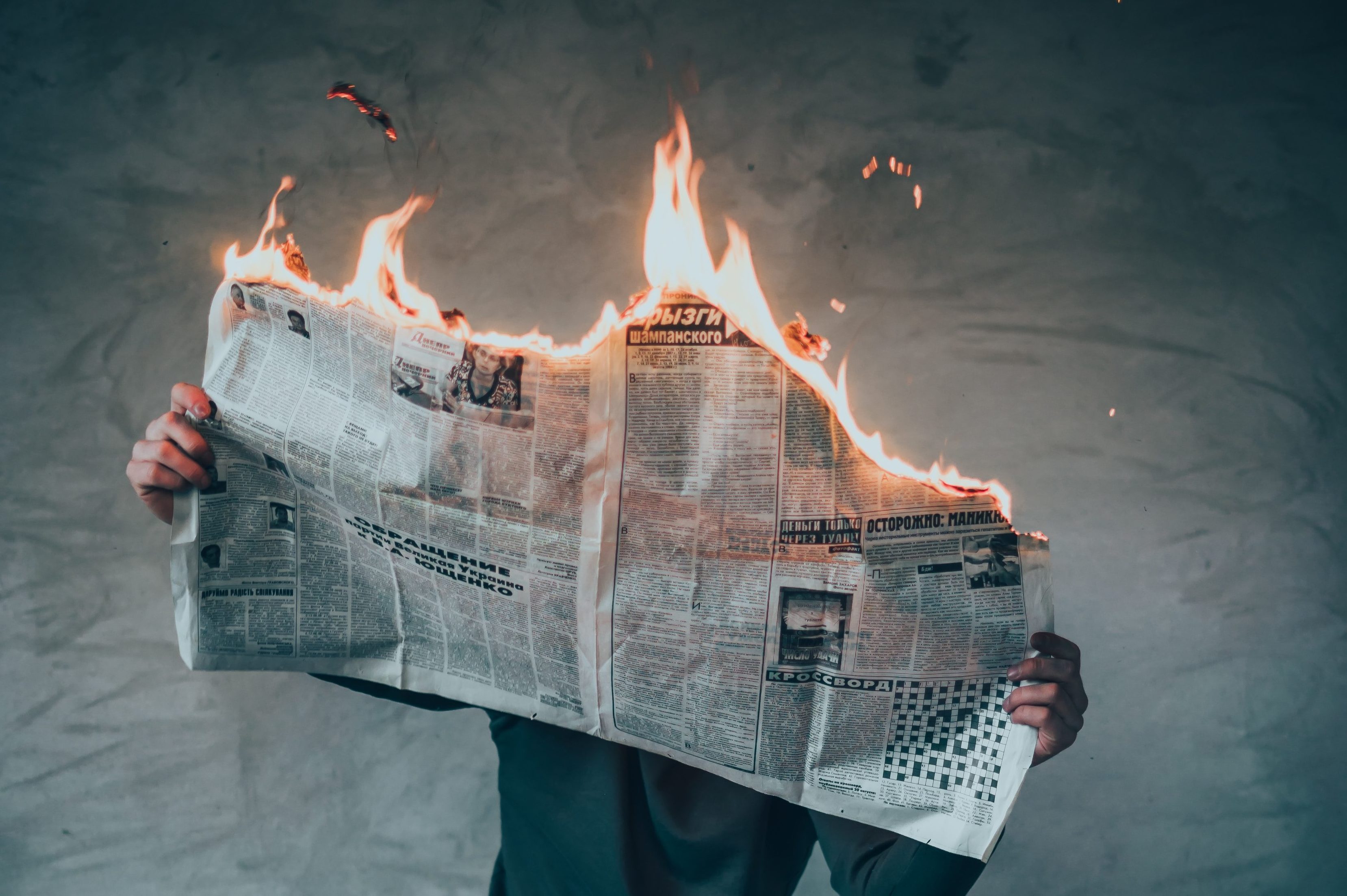 A man reads a newspaper that is on fire while standing against a wall