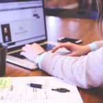 A young woman wearing a watch types on a laptop with a legal pad covered in notes on her desk