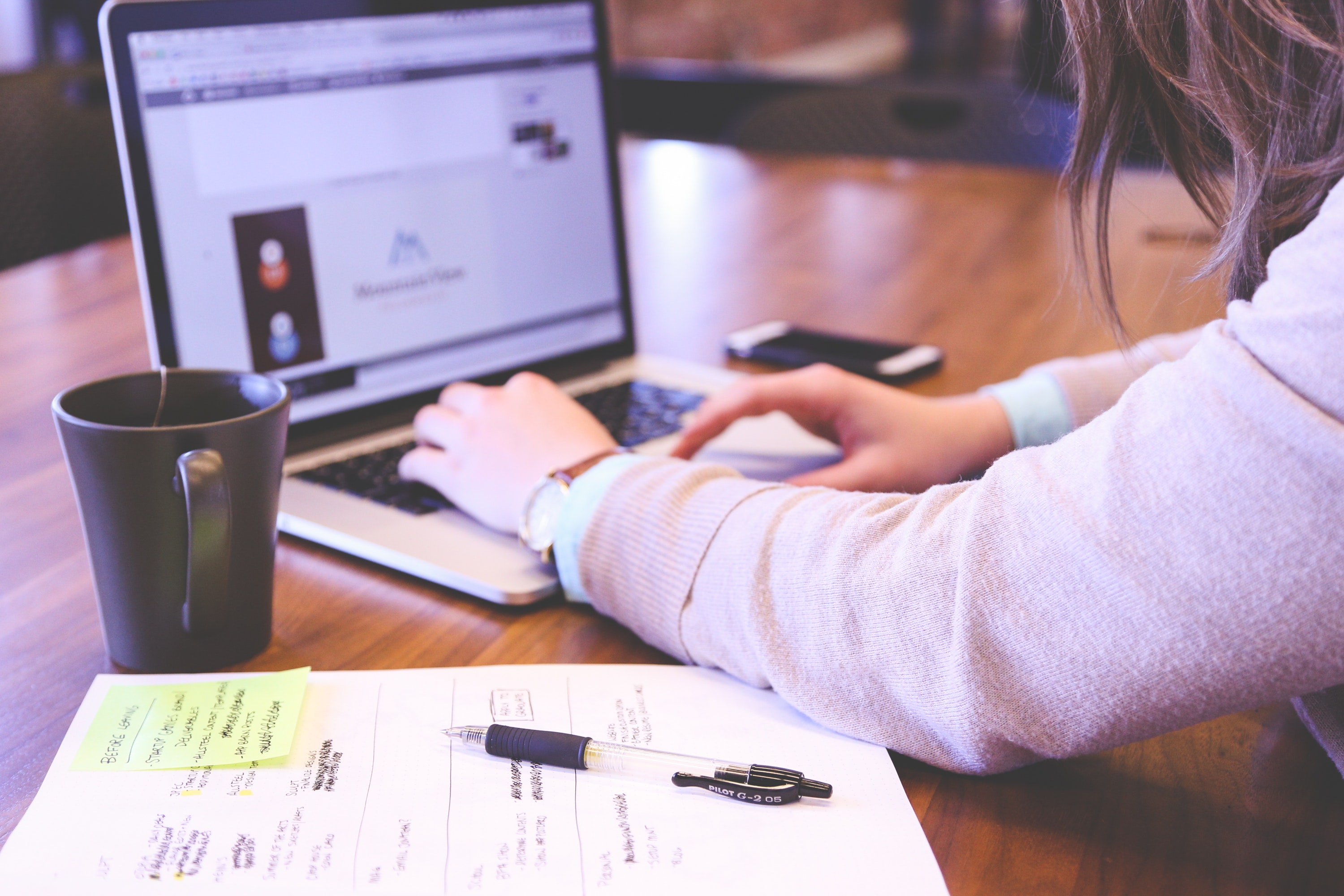 A young woman wearing a watch types on a laptop with a legal pad covered in notes on her desk