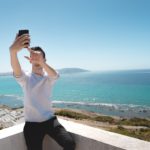 A man takes a selfie on the roof of a white building with a sunny beach in the background