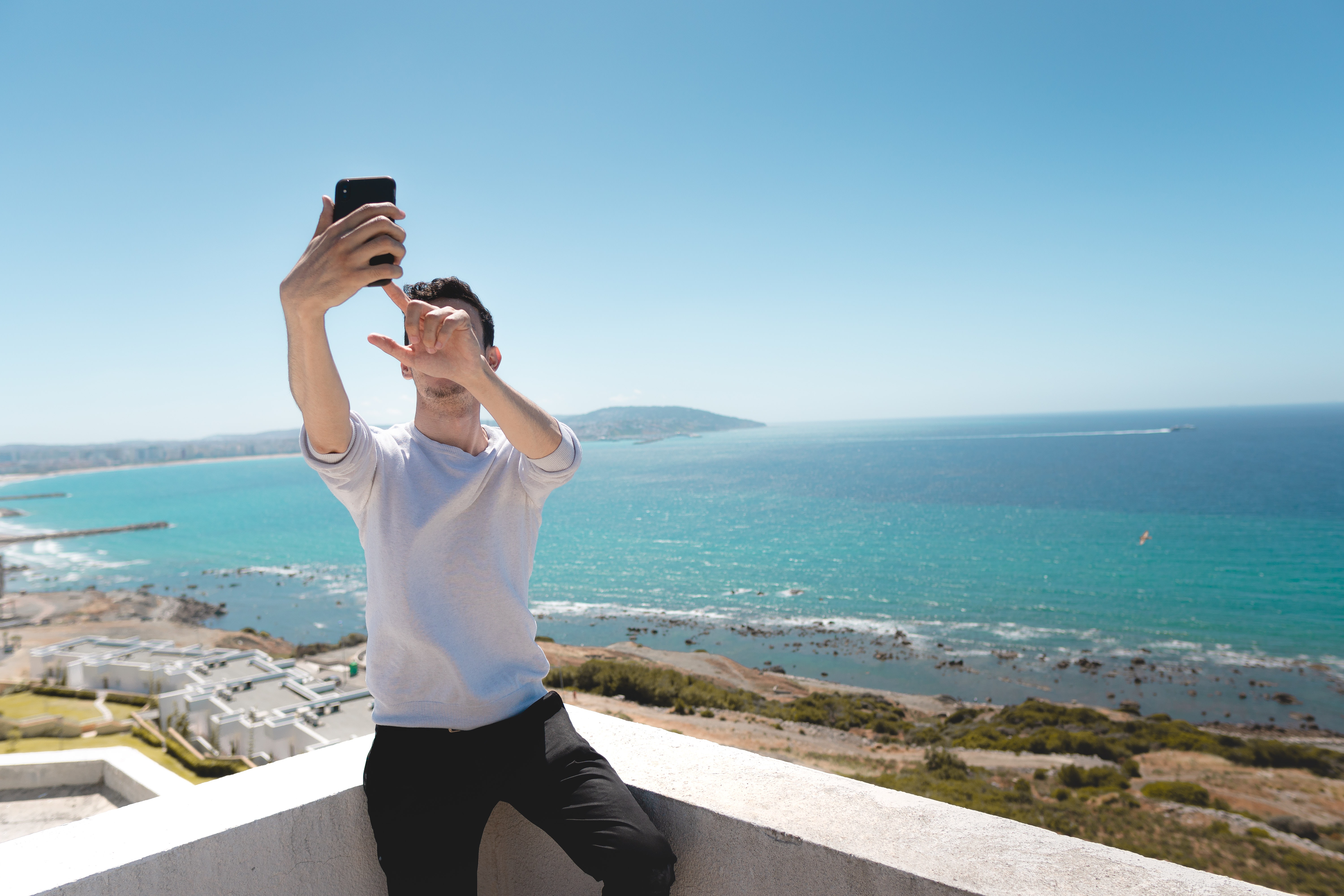 A man takes a selfie on the roof of a white building with a sunny beach in the background